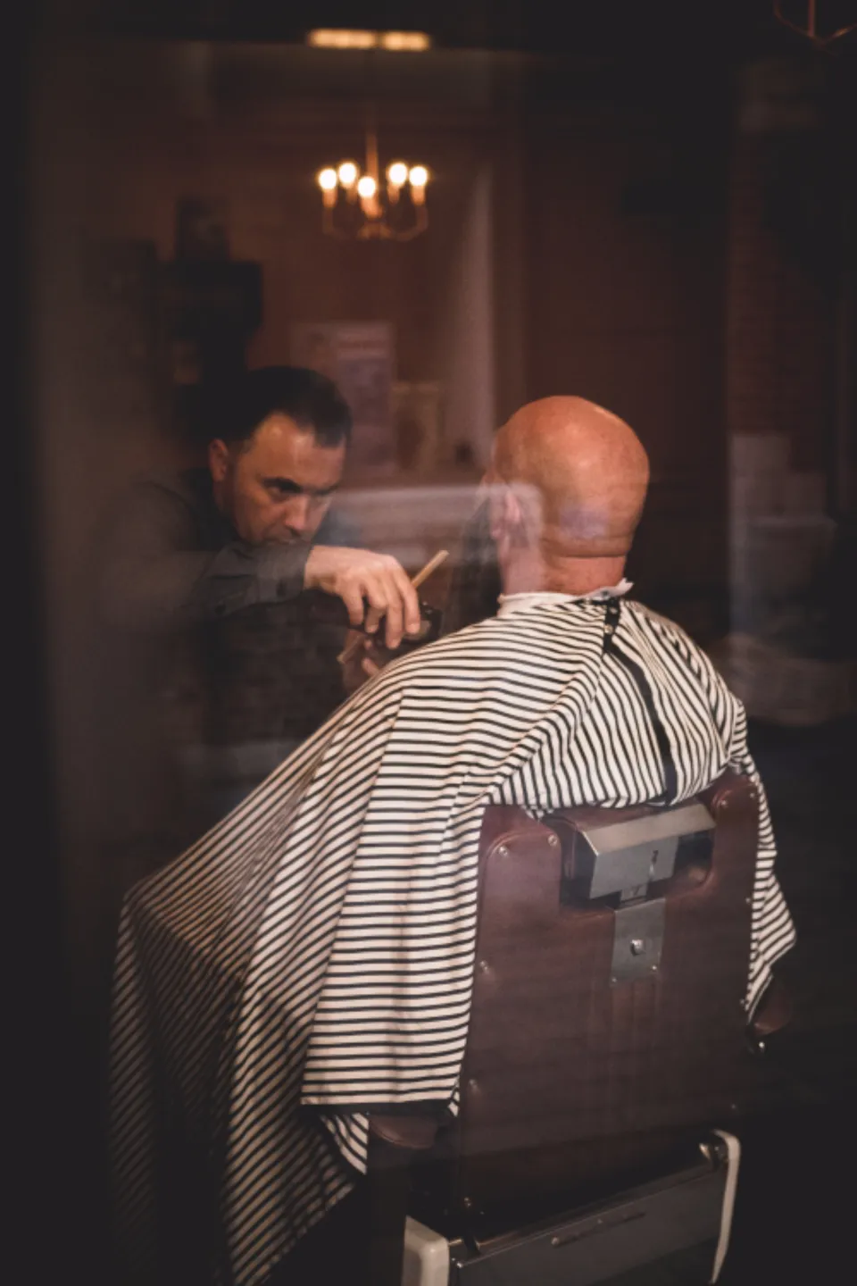 Barbier au travail dans un salon tamisé, client assis sous une cape rayée