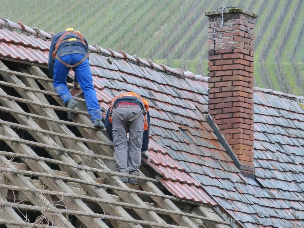 Deux couvreurs en harnais sur une toiture en tuiles en cours de rénovation, près d'une cheminée en briques
